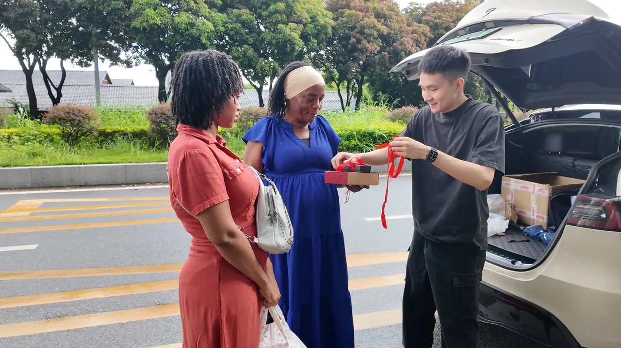 Three people smile and look at a gift box beside an open car boot.