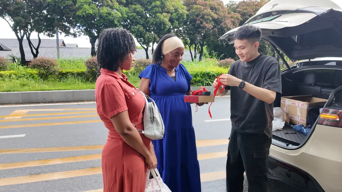 Three people look at a ribboned gift box beside an open car boot.